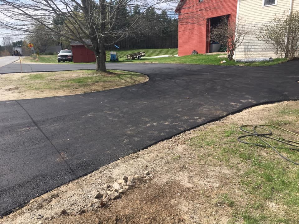A black driveway leading to a house with a red barn in the background.