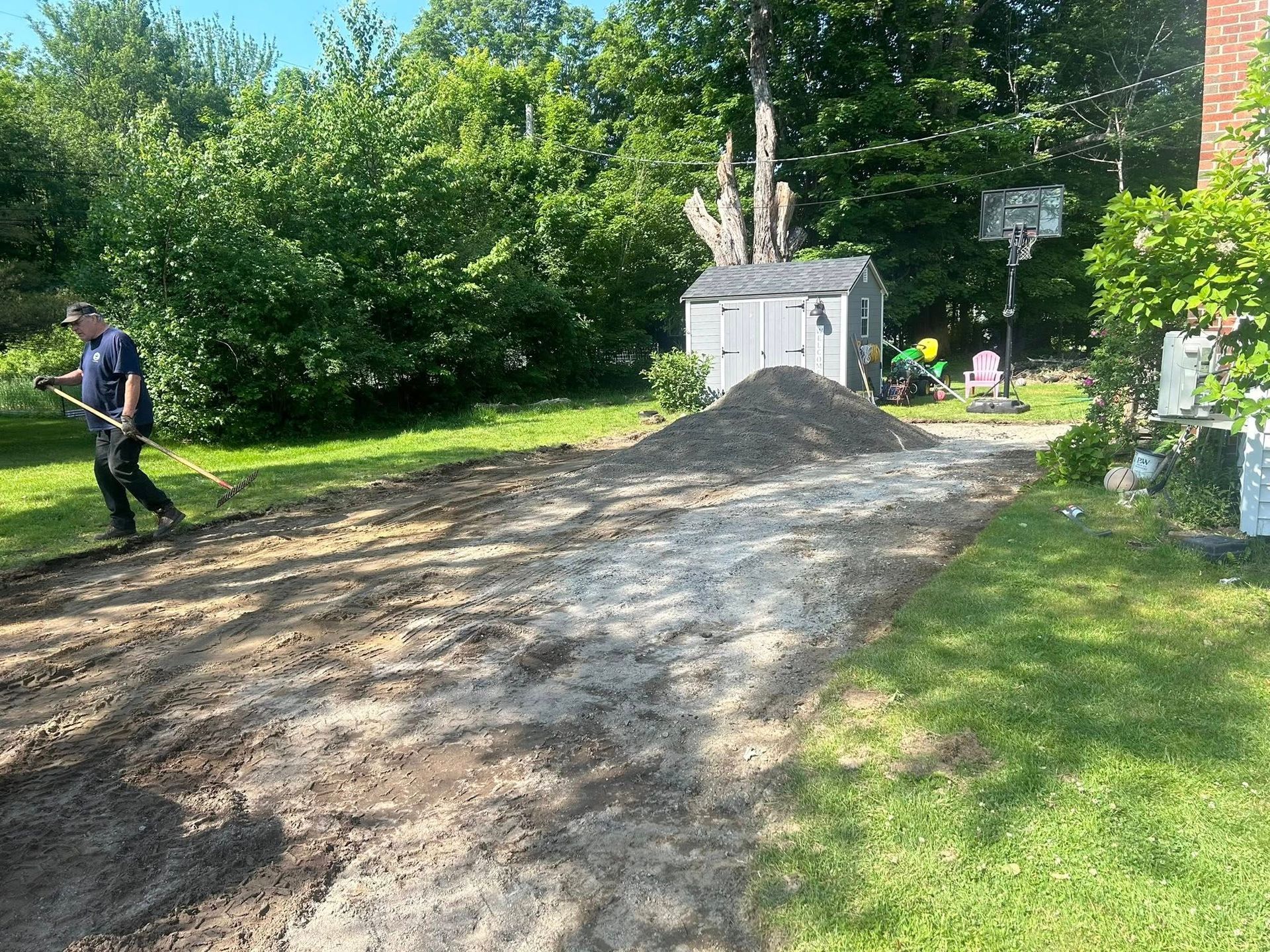 A man is raking dirt in a driveway next to a shed.