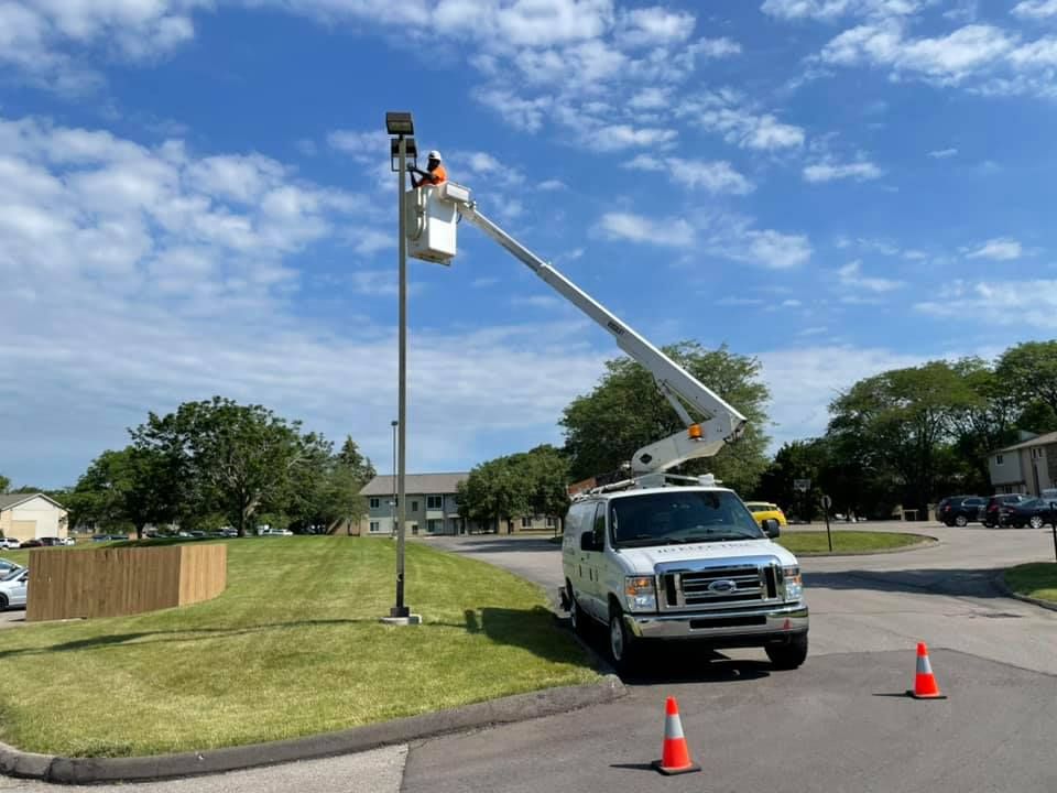 Bucket truck repairing a tall pole near a grassy roadside, with orange cones under a blue sky.