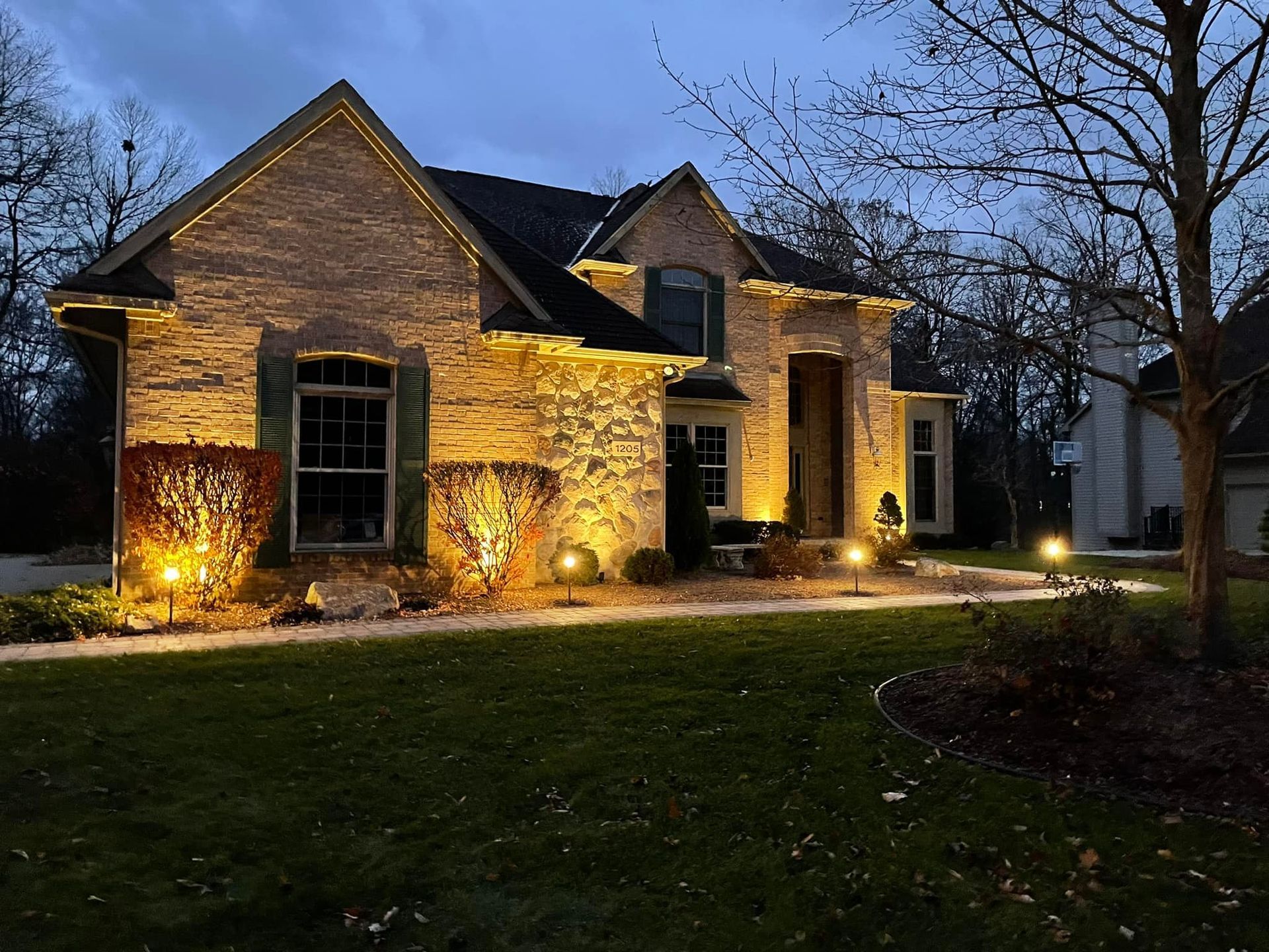 Brick house at dusk with warm exterior lights illuminating the stone facade and front yard