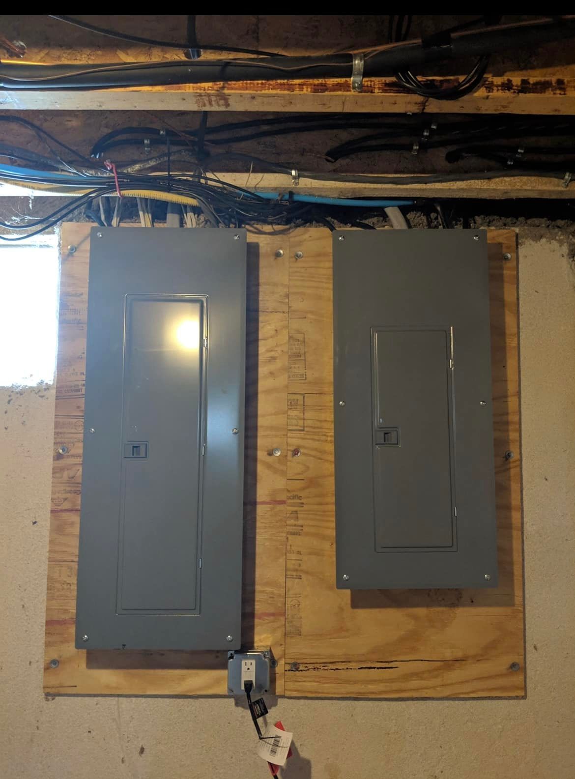 Two gray electrical panels mounted on plywood in a basement ceiling area