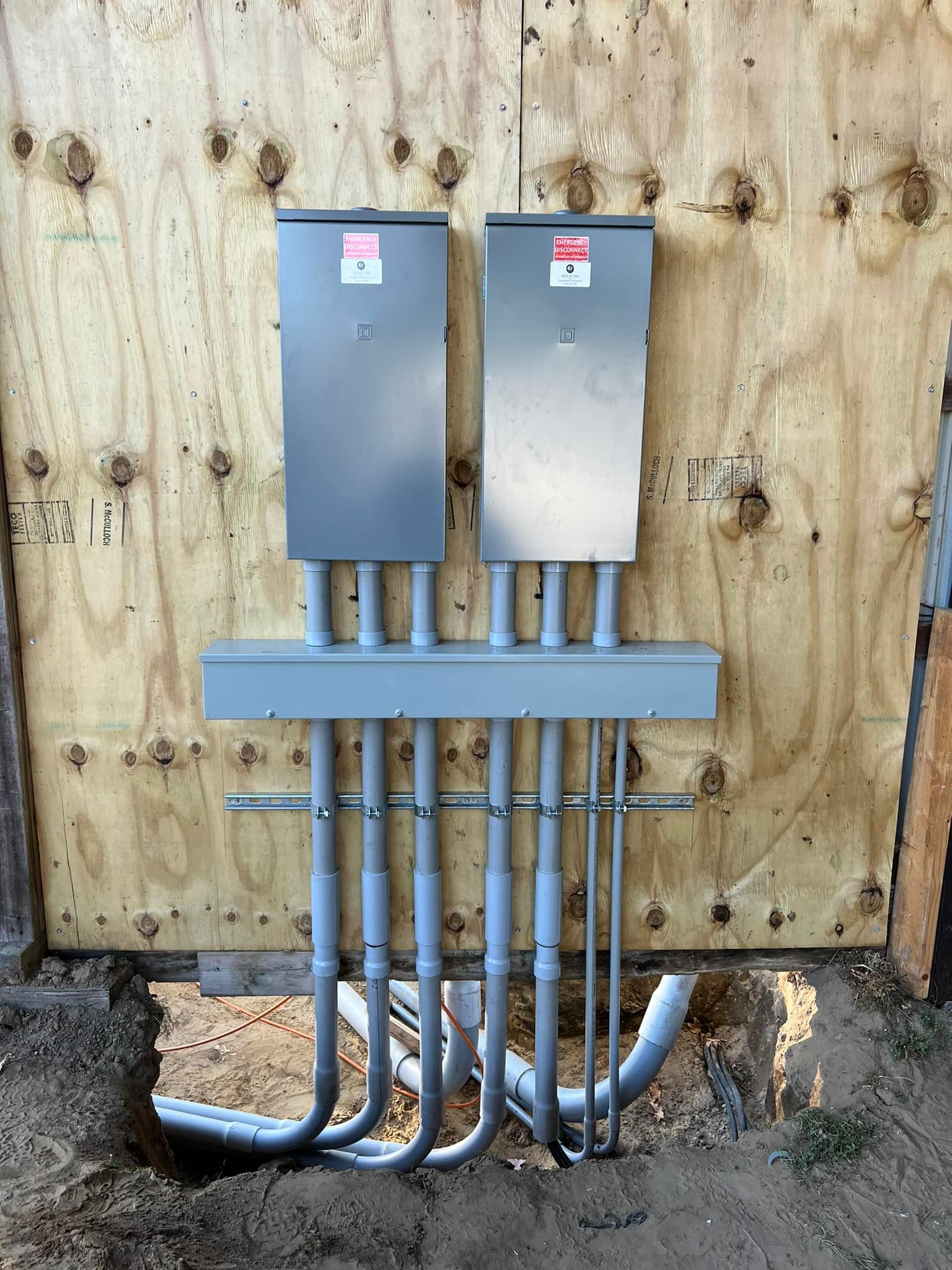 Two gray electrical panels and conduit pipes mounted on a plywood wall outdoors