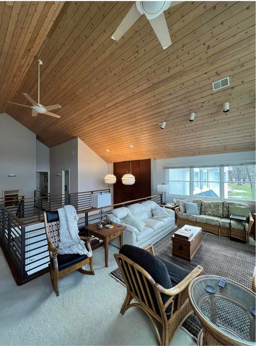 Cozy living room with wood-paneled vaulted ceiling, wicker chairs, sofa, and large windows overlooking water.