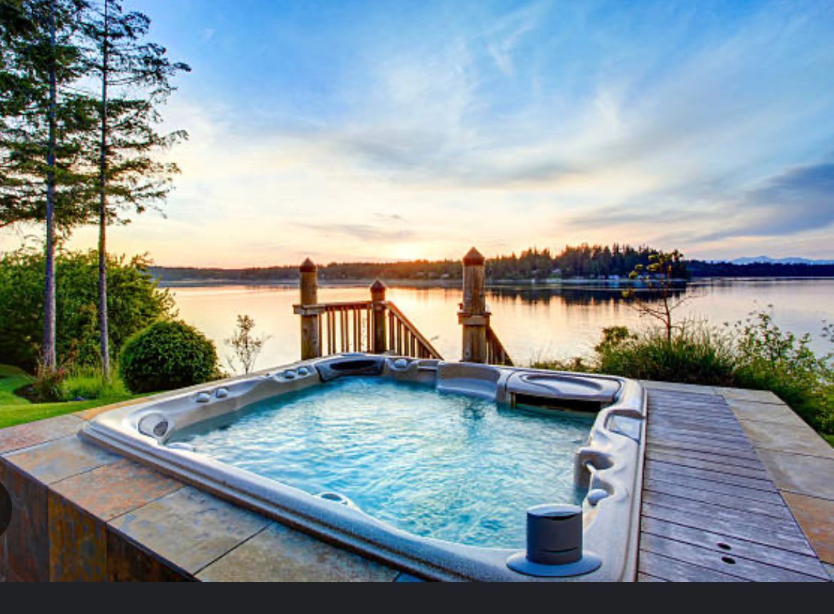 Hot tub on a wooden deck overlooking a calm lake at sunset, with trees and a dock in the background