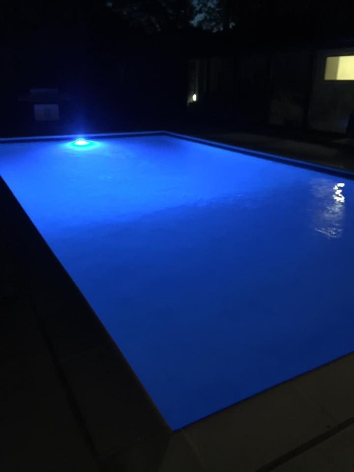 Blue-lit swimming pool at night with dark surroundings and a lit window in the background