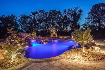 Lit backyard pool glowing blue at night, surrounded by stone patio and landscape lighting.