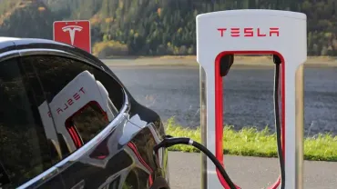Tesla car charging at a Supercharger station outdoors, with a plug connected to the rear port.