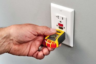 Hand holding a plug-in circuit tester at a wall outlet with red indicators lit