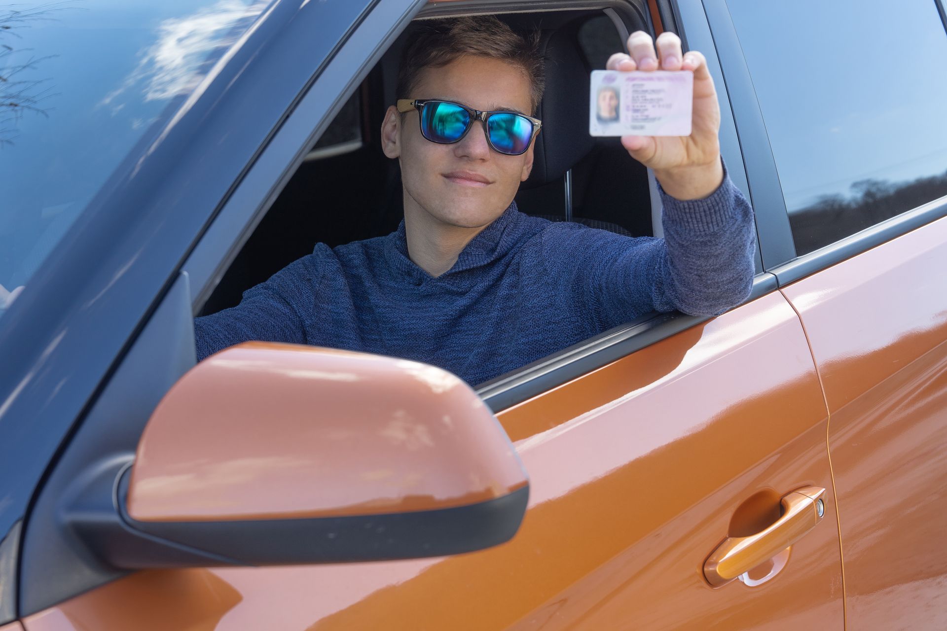 Teen driver sitting in car, smiling and holding up new driver's license.