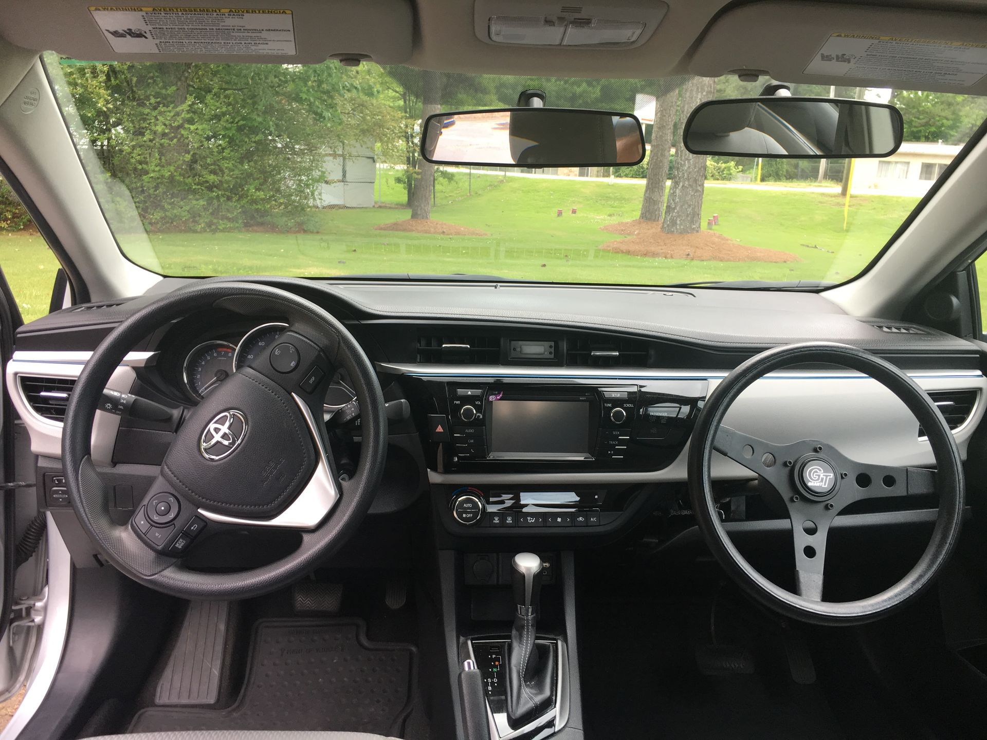 Car interior with right-hand steering wheel and manual gear shift, parked beside a green lawn.