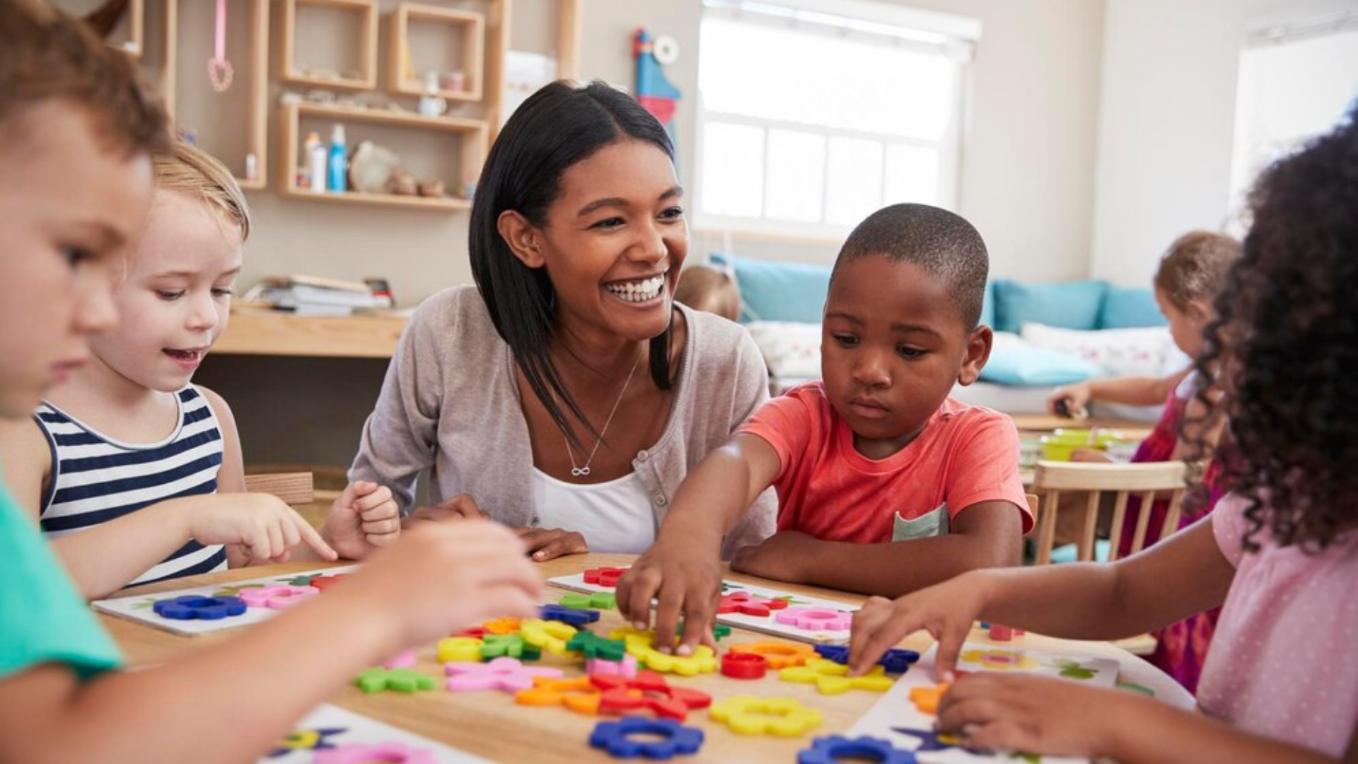 Teacher smiles, interacting with children at a table filled with colorful objects in a bright classroom.