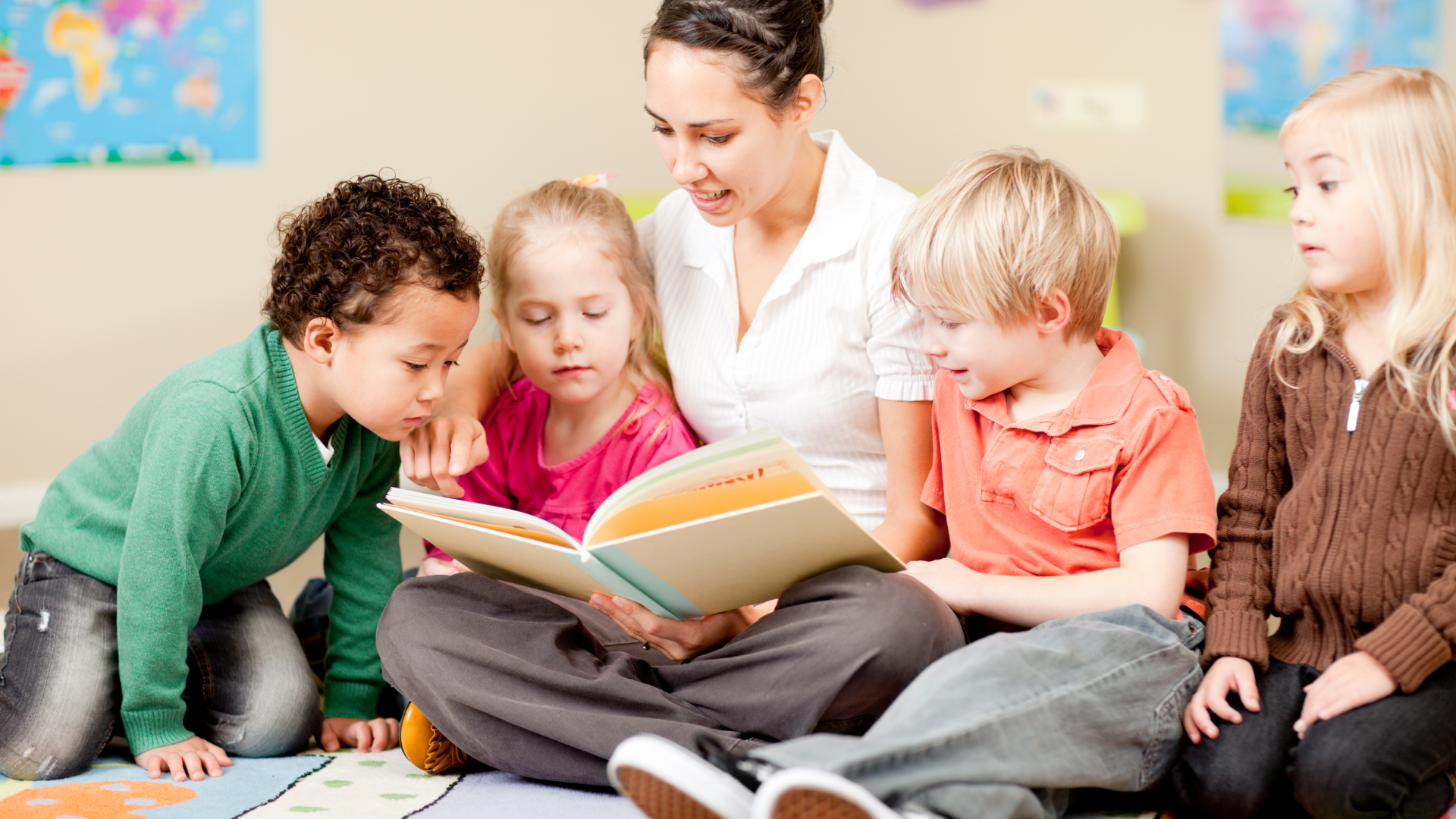 Teacher reads a book to four children seated on a floor; light-colored room.