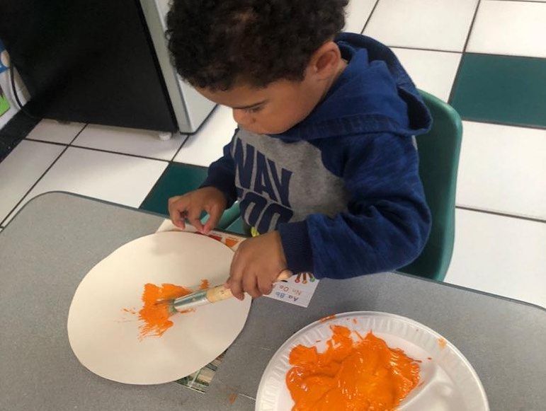 A young boy wearing a navy shirt is painting a paper plate with orange paint