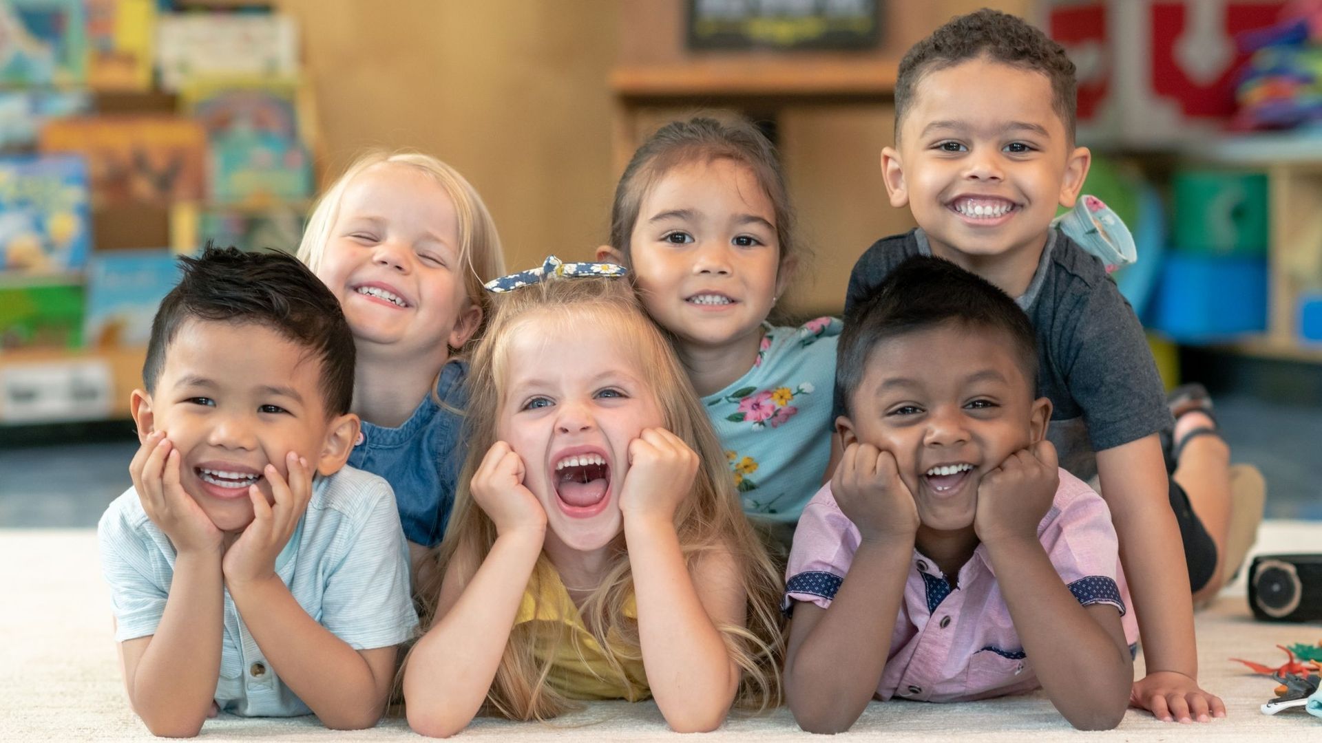 Group of children smiling and laughing, lying on a rug in a colorful classroom.