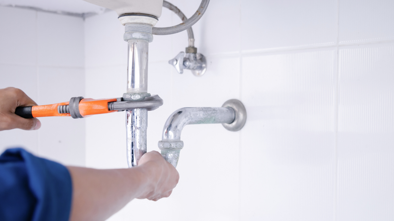 Plumber using a wrench on a sink drain pipe; white tile wall background.