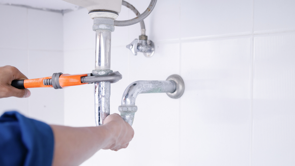Plumber using wrench on chrome pipes under a sink; white tiled wall.