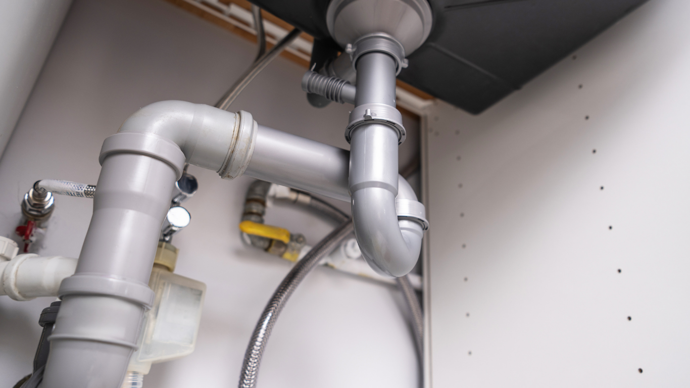 Plumbing pipes under a white sink, including a curved drainpipe, connecting to a waste disposal and water supply.