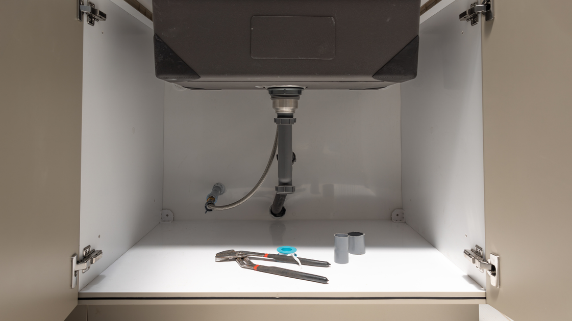 Undersink plumbing: grey drain pipes, flexible hose, garbage disposal, and wooden cabinet interior.