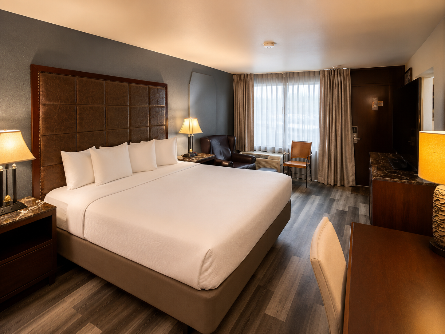 Hotel room with a large white bed, warm lamps, desk, curtains, and city-view window