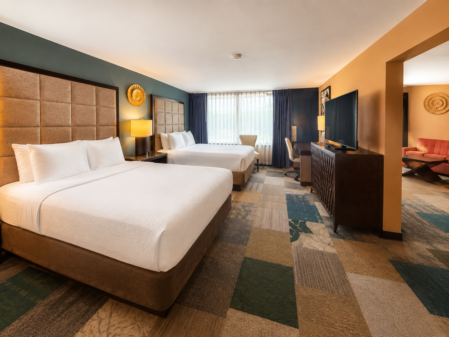Modern hotel room with two beds, patterned carpet, TV, and window seating area