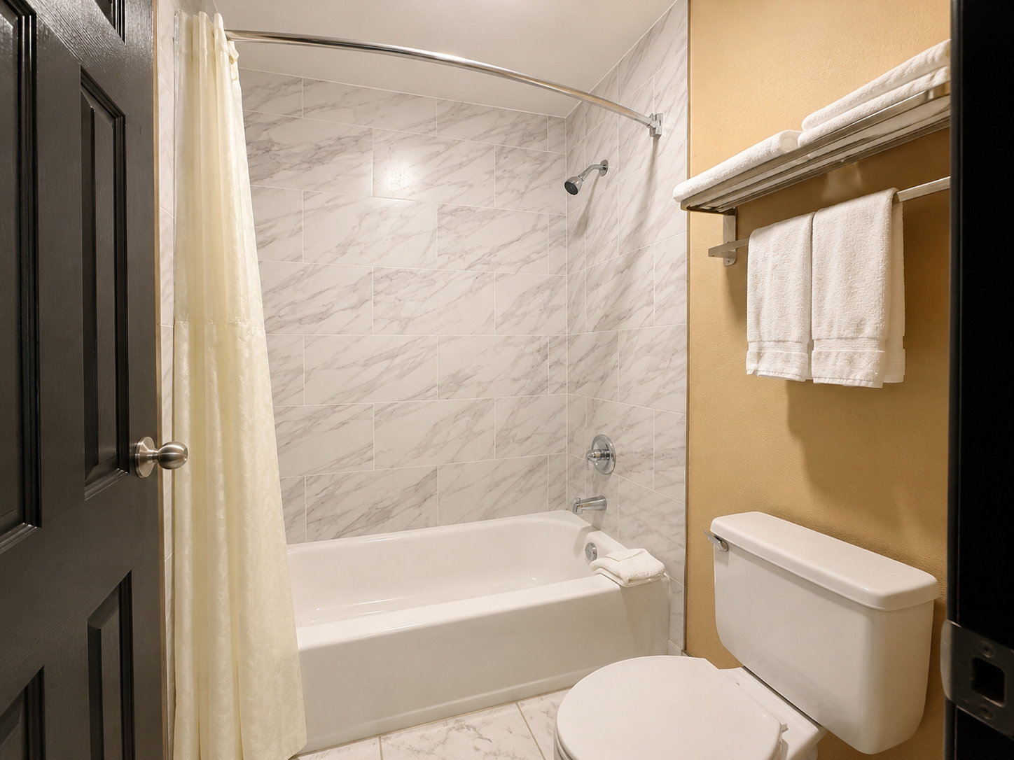 Small bathroom with tub-shower, marble walls, beige walls, toilet, towel rack, and shower curtain.