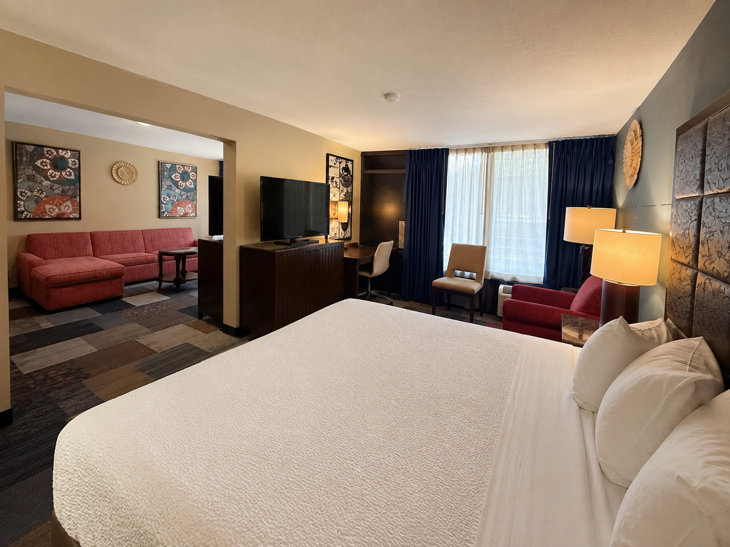 Hotel room with large bed, sofa, TV, and window seating in warm lighting