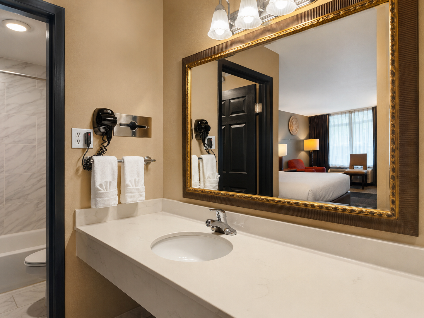 Hotel bathroom vanity with large gold-framed mirror, sink, and towels beside a wall-mounted hair dryer