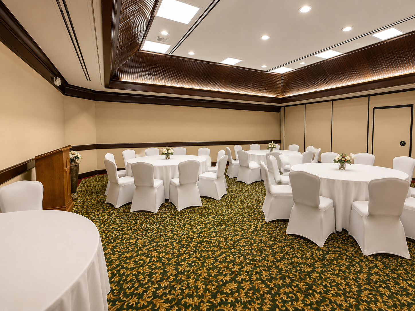 Empty banquet room with round tables, white chair covers, and floral centerpieces on patterned carpet