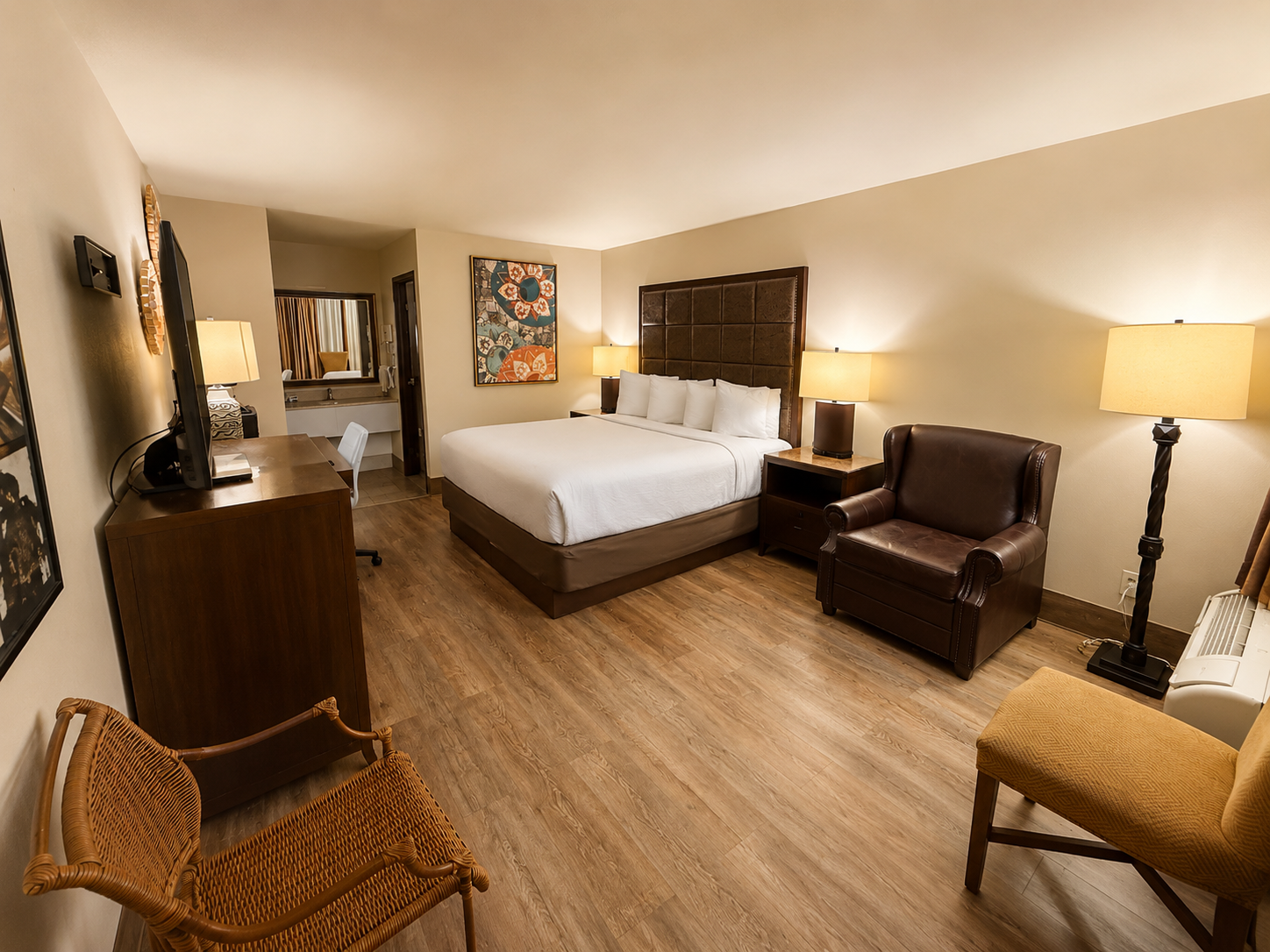 Cozy hotel room with king bed, armchair, floor lamps, wood flooring, and warm beige walls