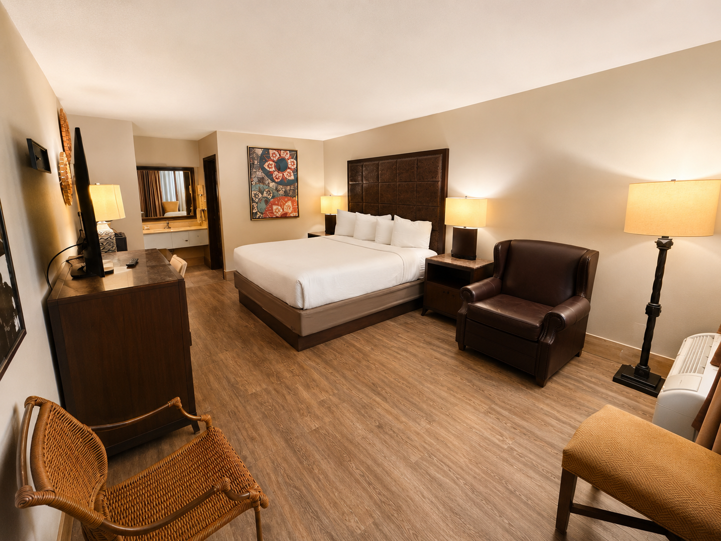 Modern hotel room with king bed, leather chair, lamps, wicker chair, and wood flooring
