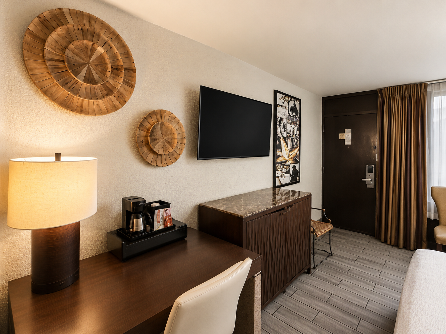 Hotel room with wall-mounted TV, desk lamp, woven wall decor, and a closed door.