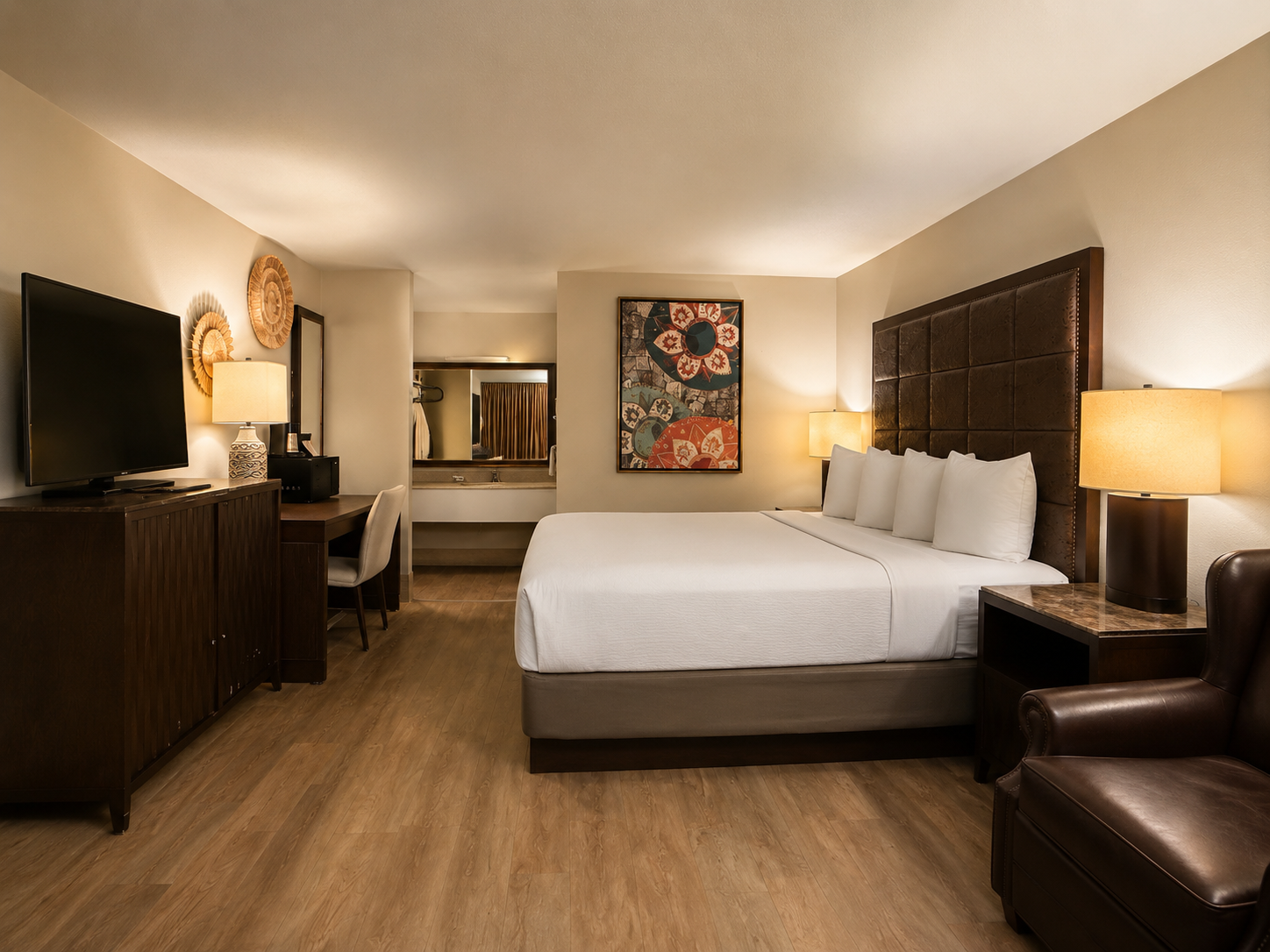 Modern hotel room with king bed, warm lighting, TV, desk, and armchair on wood flooring