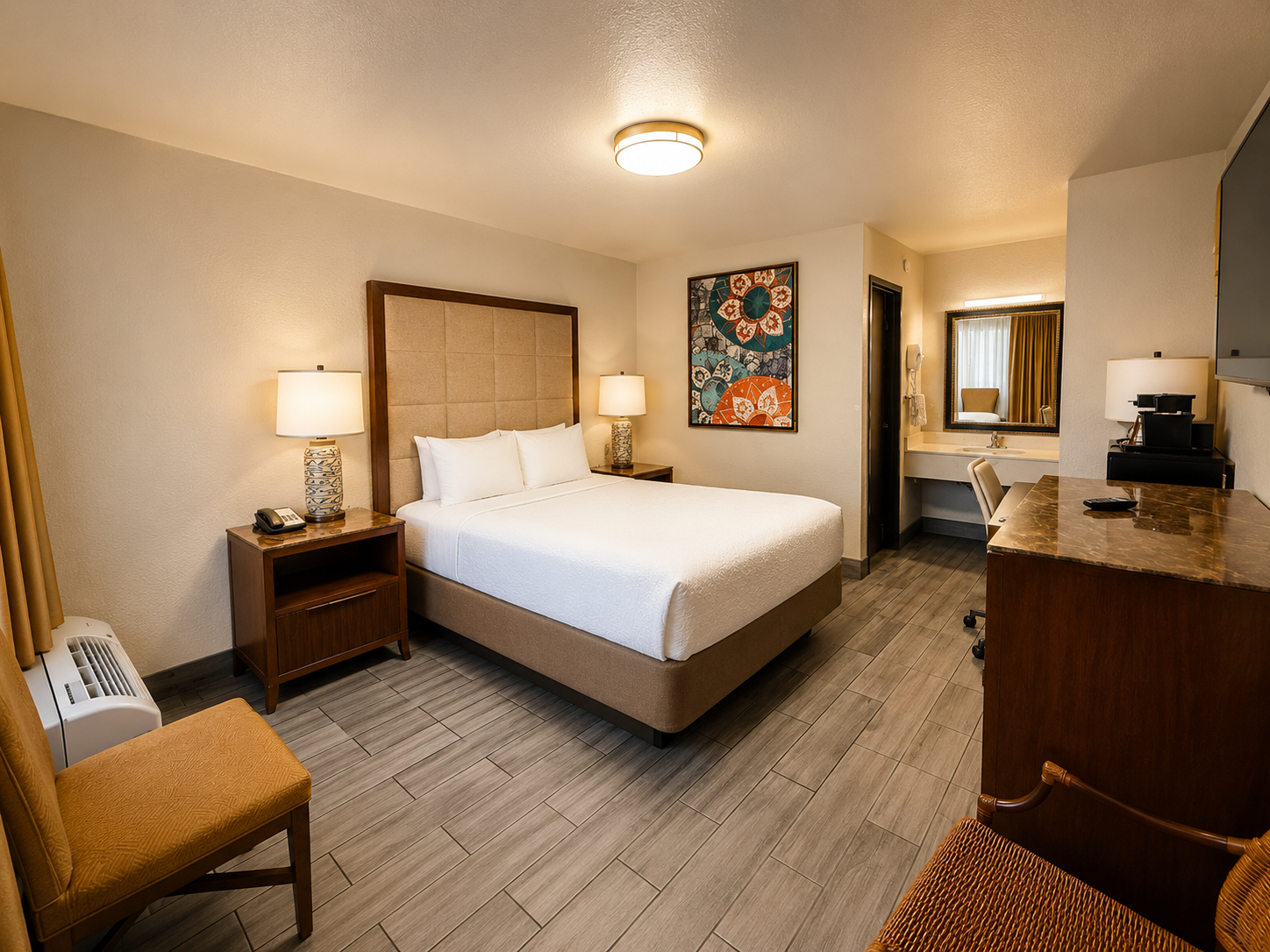 Hotel room with a neatly made bed, bedside lamps, desk, chair, and a bathroom visible through an open doorway.