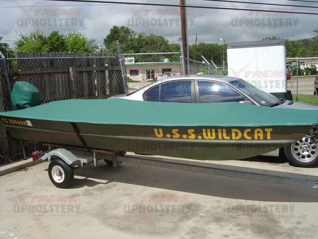 Green boat on trailer with “U.S.S. Wildcat” painted on the side, parked beside a car outdoors