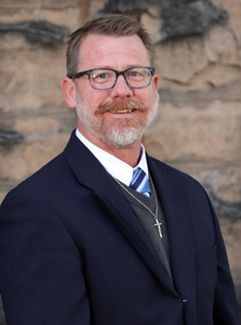 A man wearing glasses and a blue sweater is smiling in front of a stone wall.