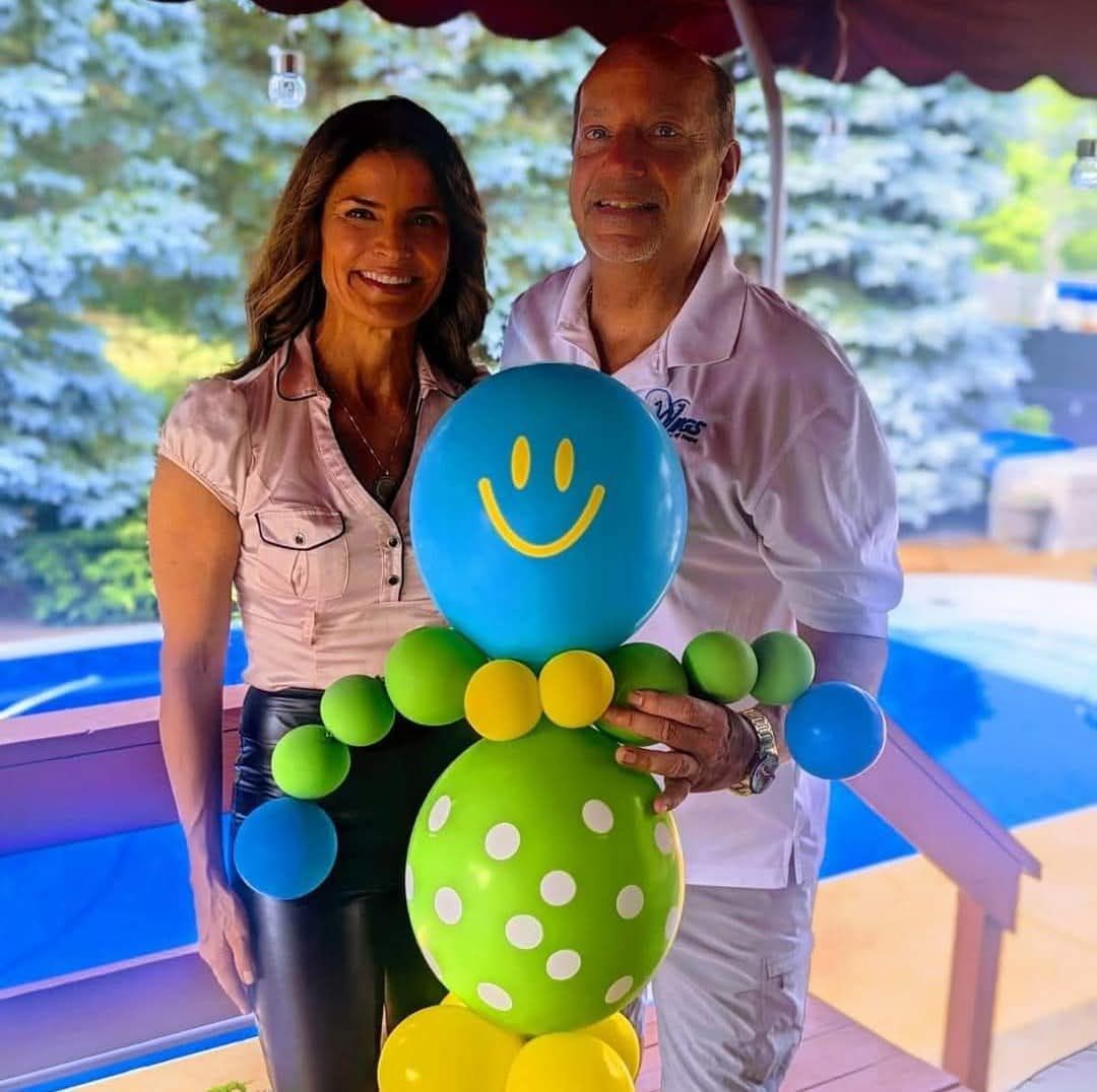 Two people posing with a colorful balloon figure on a balcony, with a scenic outdoor view behind them.