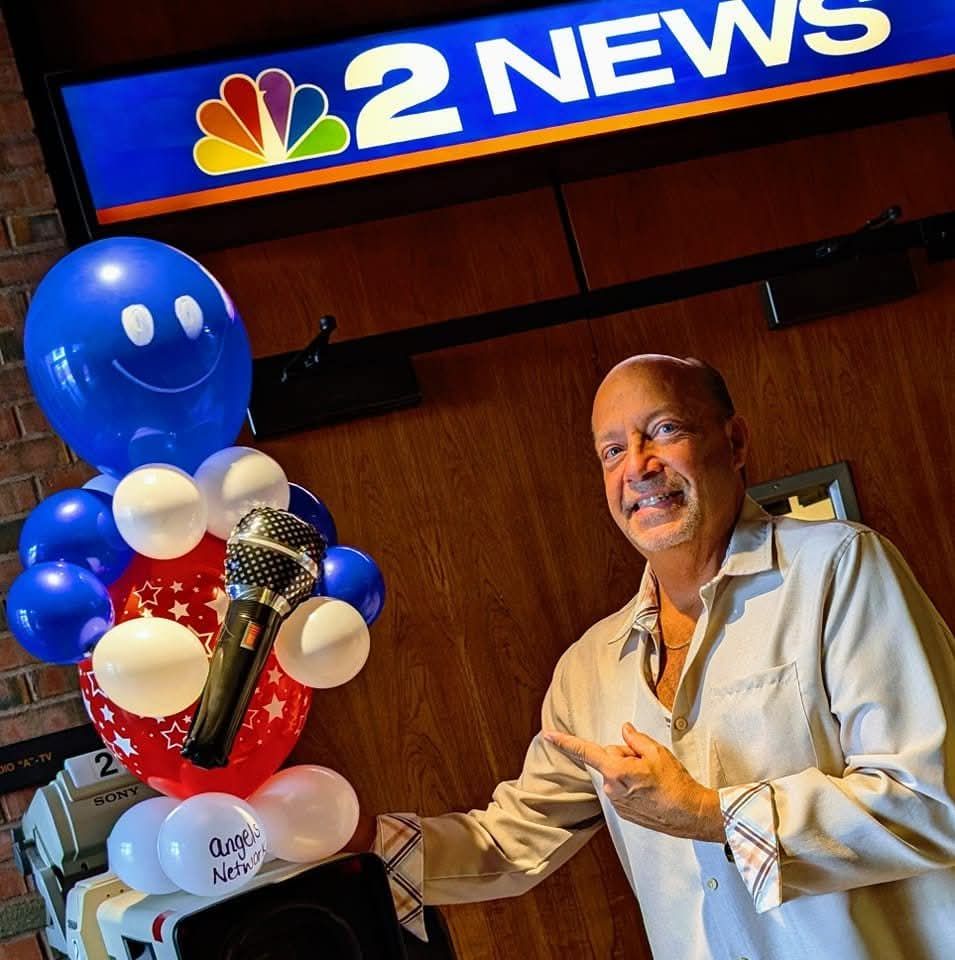 Man smiling beside NBC 2 News balloon display and microphone in a studio.