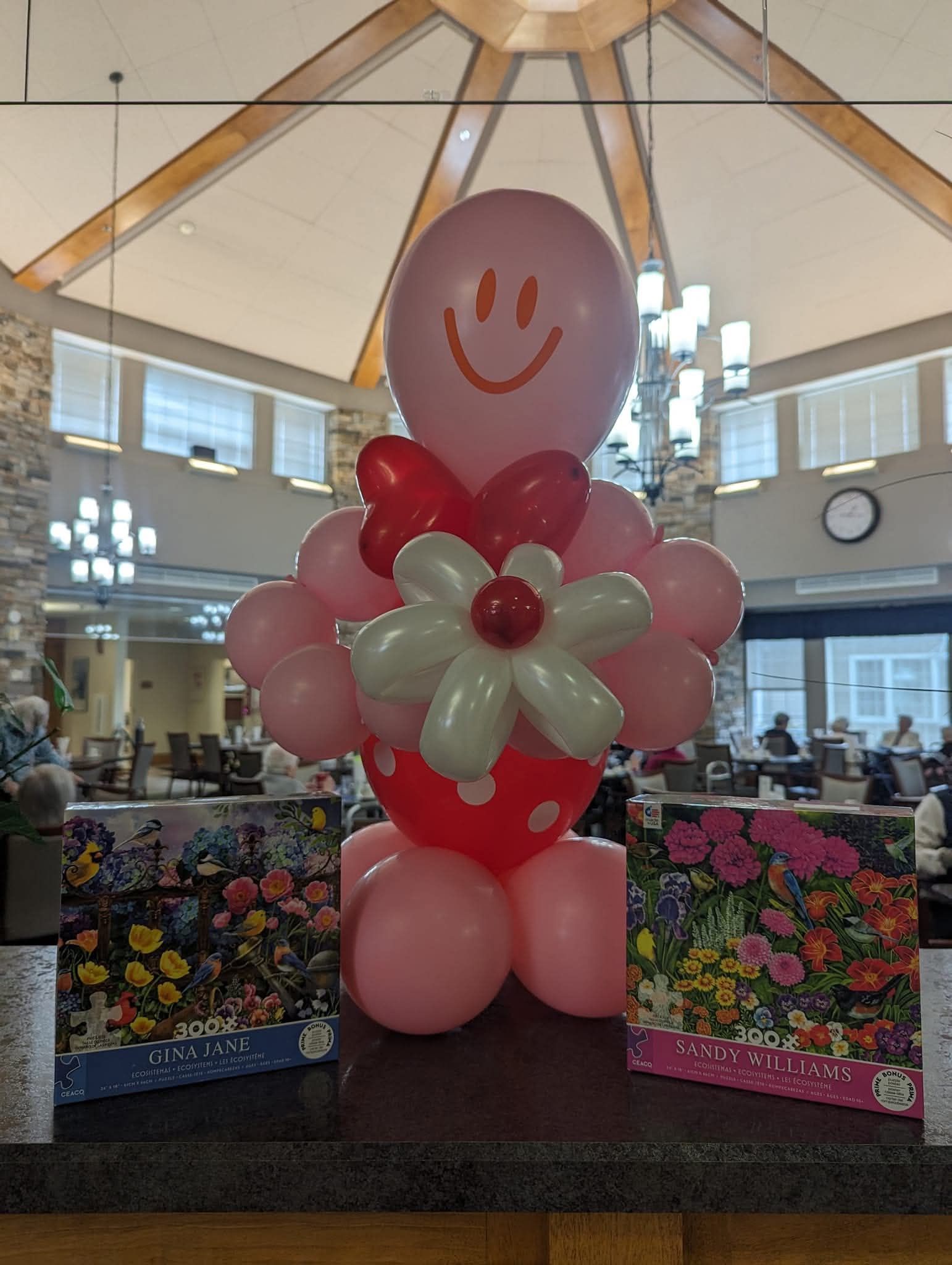 Pink balloon flower arrangement with smiley-face balloon in a bright indoor lobby, flanked by floral displays