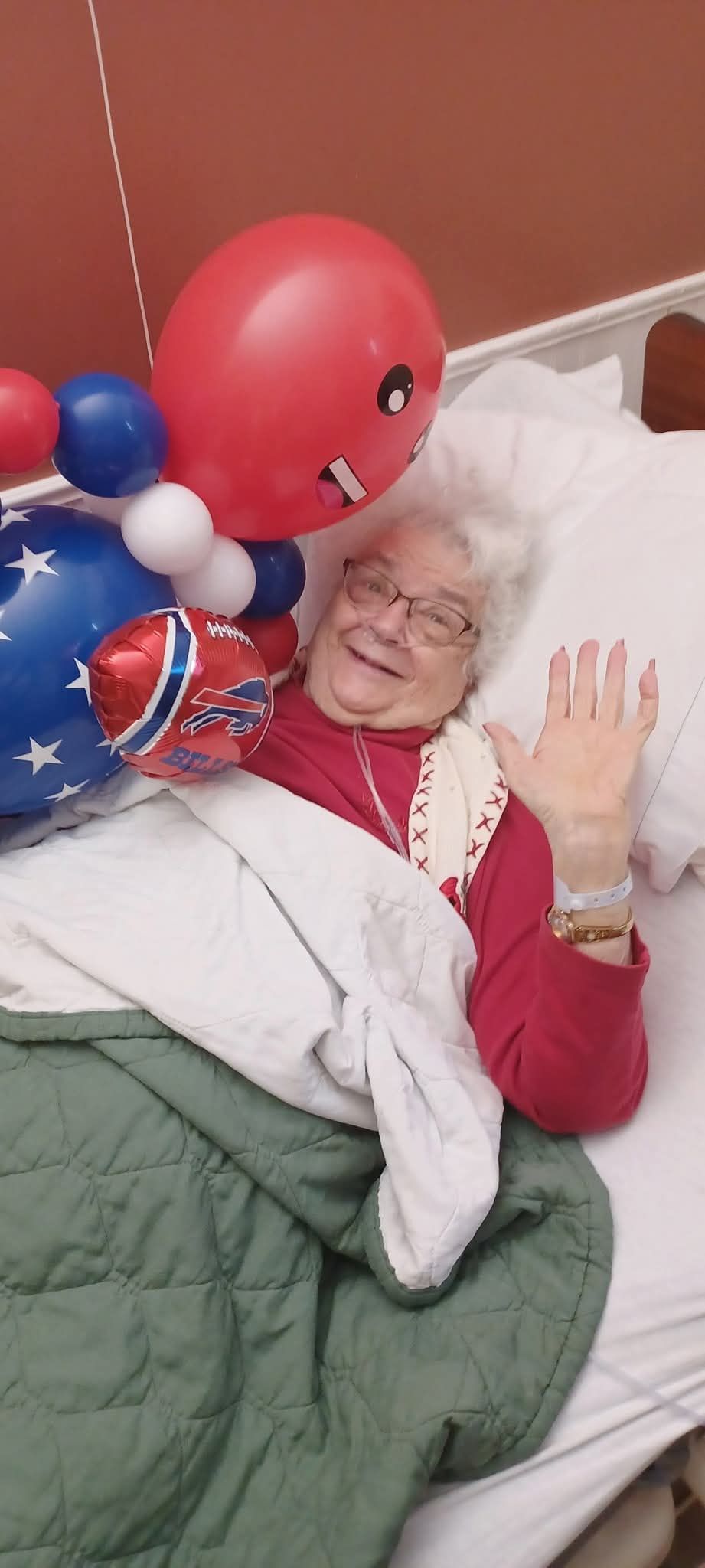 Person in bed smiling and waving, surrounded by red and blue balloons and a wrapped gift.