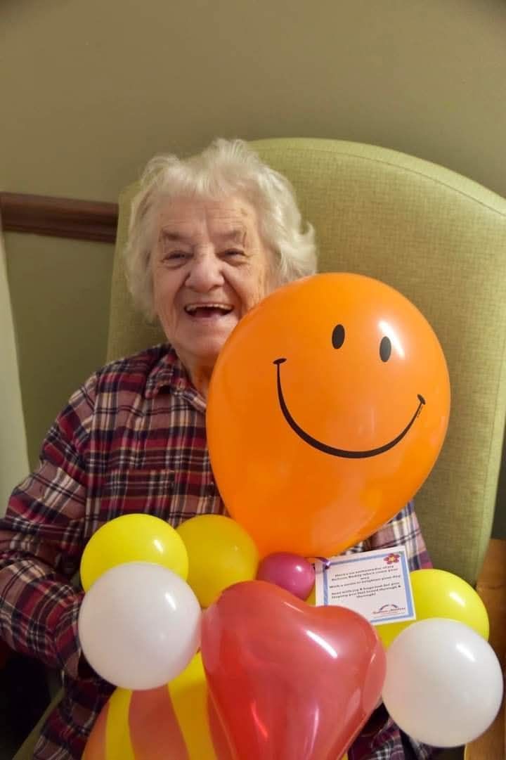 Smiling person behind orange smiley balloon, surrounded by yellow, white, and red balloons.