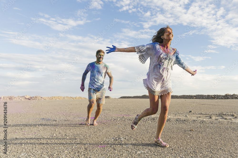 Woman running with arms open, being chased by a person, both covered in colorful powder on a beach.