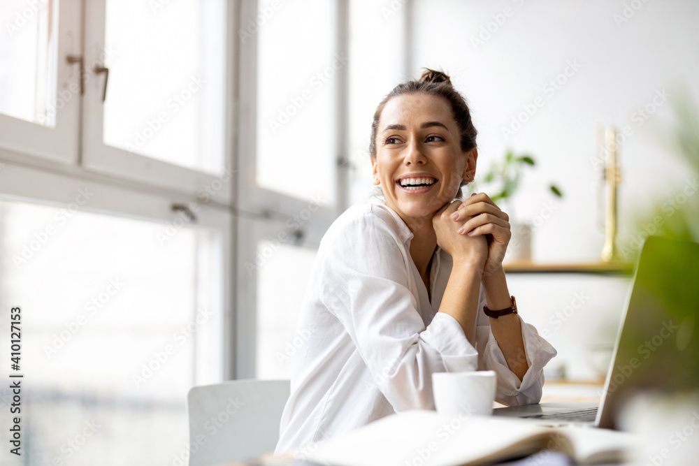 Woman smiles while sitting at a desk with a laptop and coffee mug near a window.
