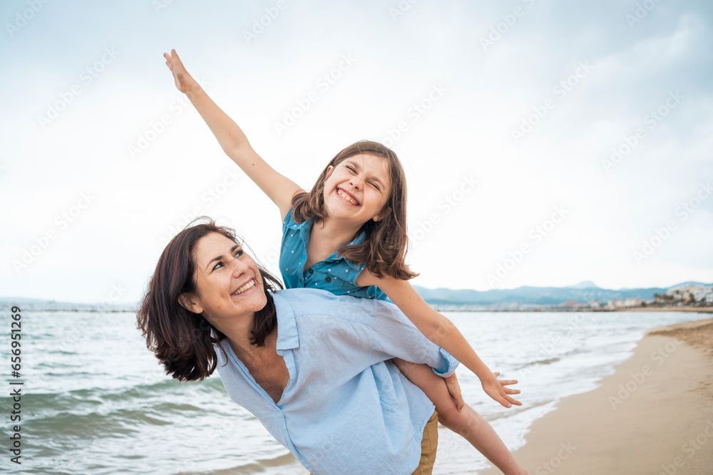 Woman giving a piggyback ride to a child on a beach, smiling. The child has arm outstretched, appearing to fly.