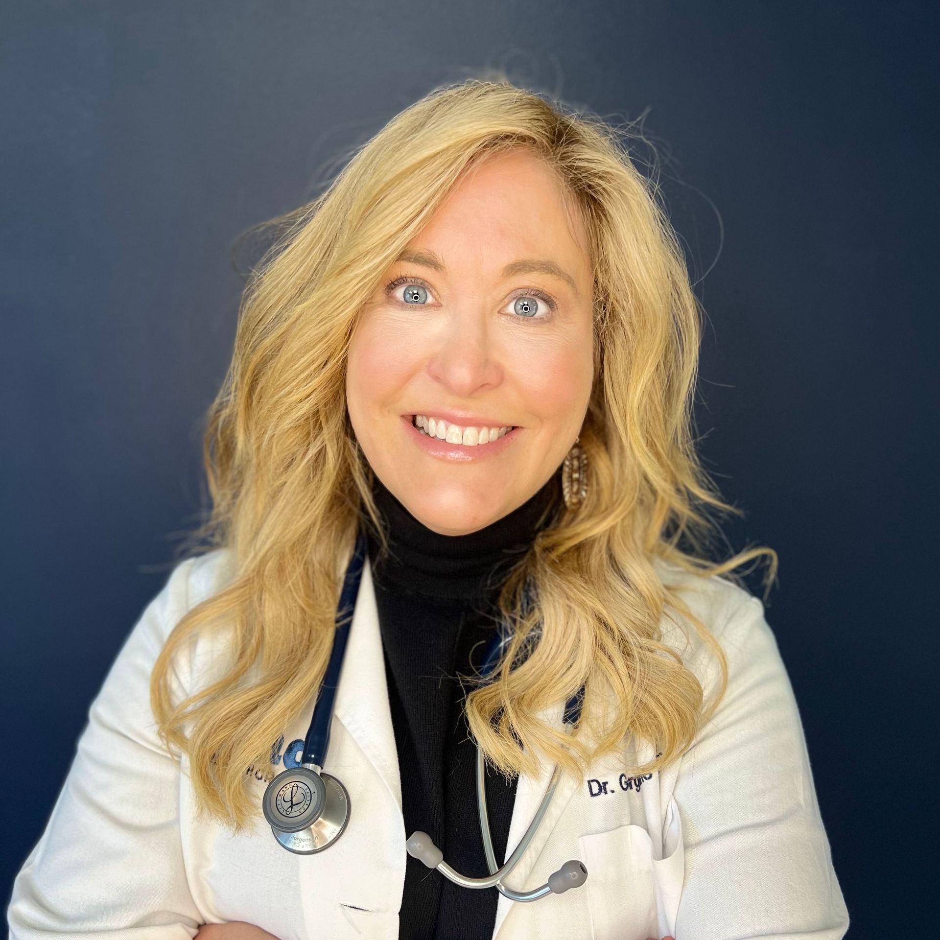 Woman in white coat with stethoscope, smiling against a dark blue background.