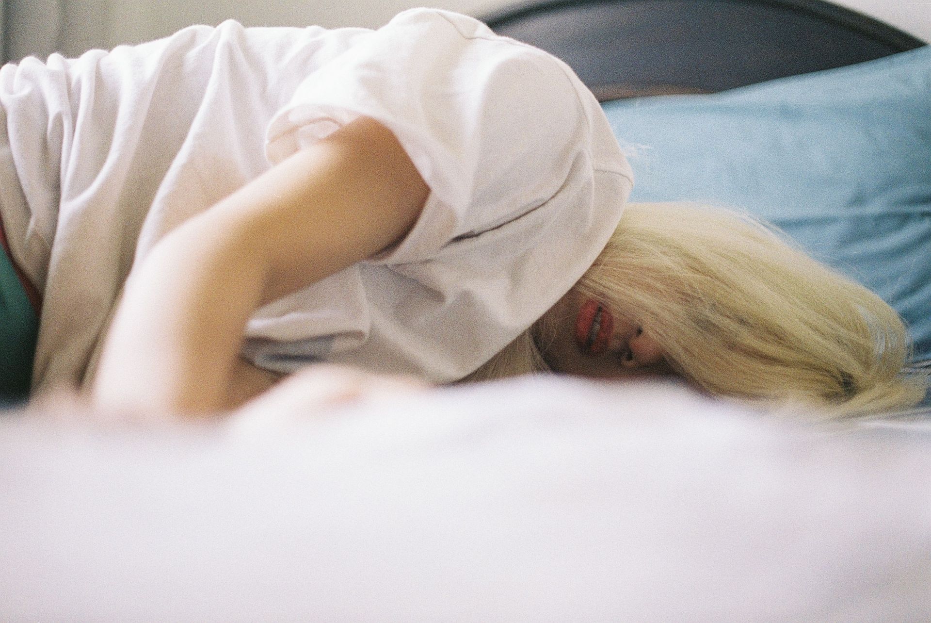 Person lying face down on a bed with a white blanket, head turned to the side, in a softly lit room