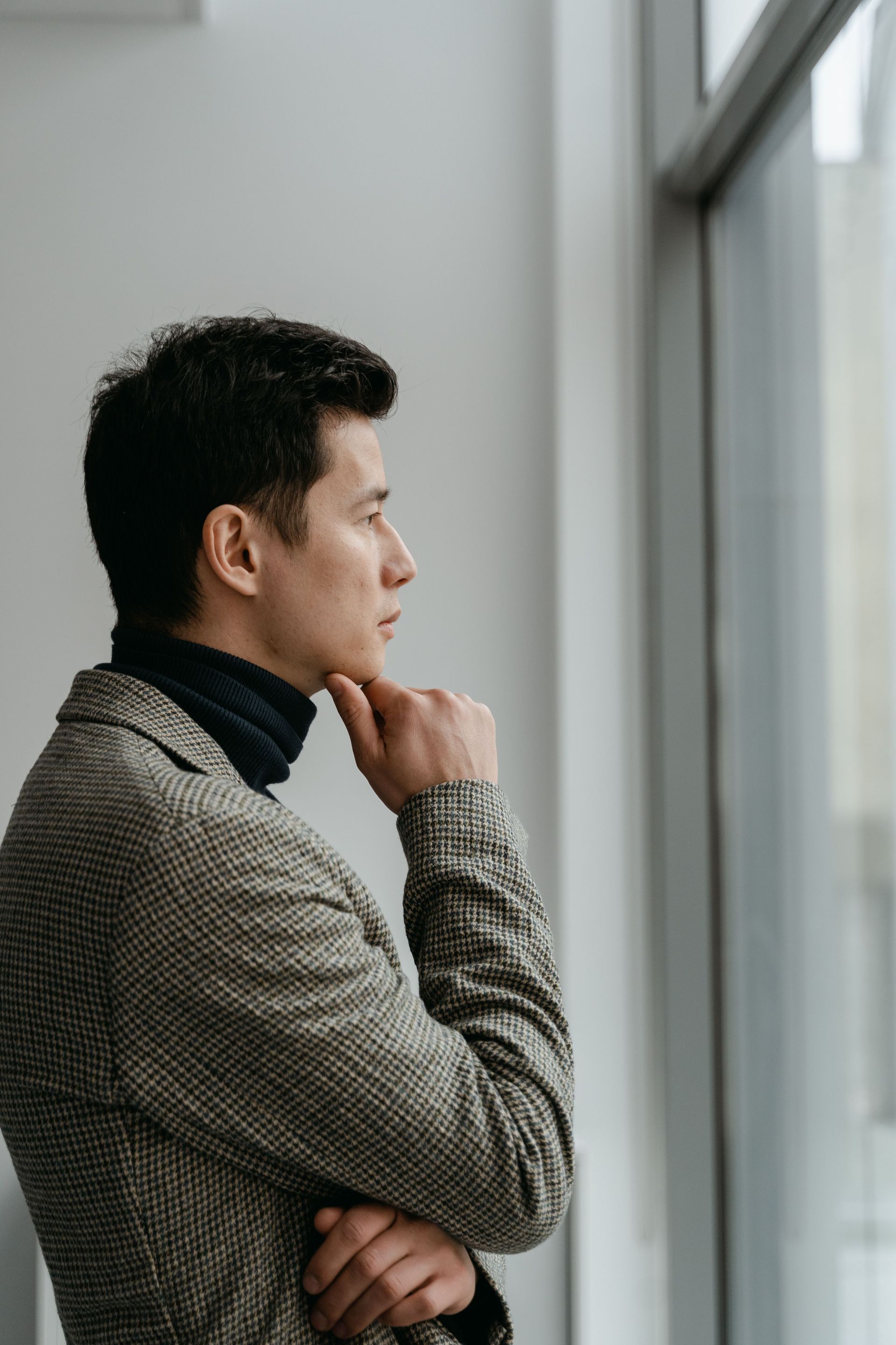 Man in a gray sweater gazes out a window with his hand on his chin, standing by a bright wall.