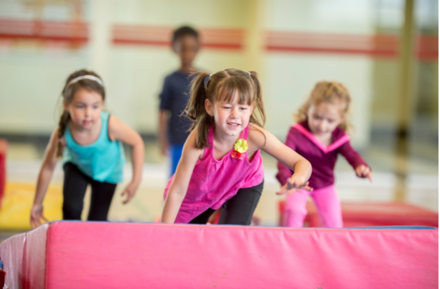 A group of young girls are jumping over a pink mat in a gym.