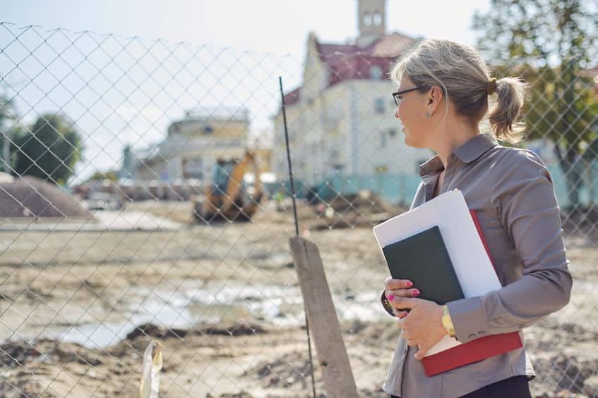 Woman holding folders at a construction site, looking left behind a chain-link fence