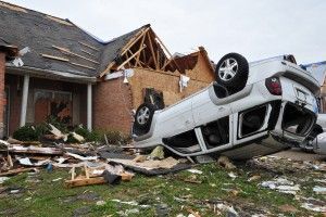 White SUV overturned near a damaged house, debris scattered on the ground.