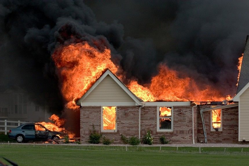 House engulfed in flames with thick black smoke, a car parked nearby, and a lawn in front